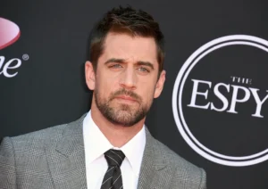 Aaron Rodgers in a grey checkered suit and striped tie at the ESPY Awards red carpet, posing in front of the ESPY logo backdrop.